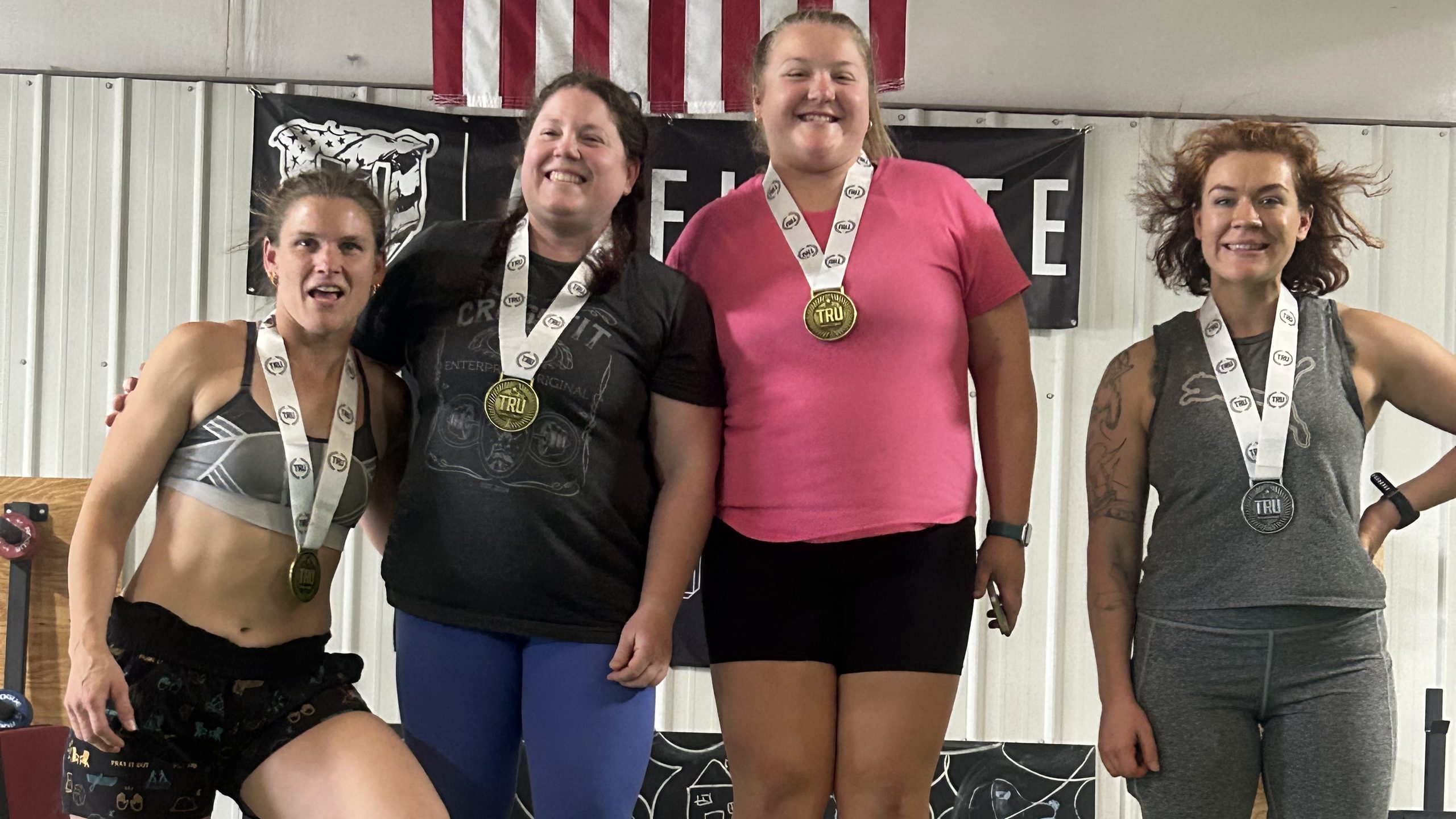 Four smiling women stand side by side wearing medals on white ribbons, with an American flag and a black wall banner behind them.
