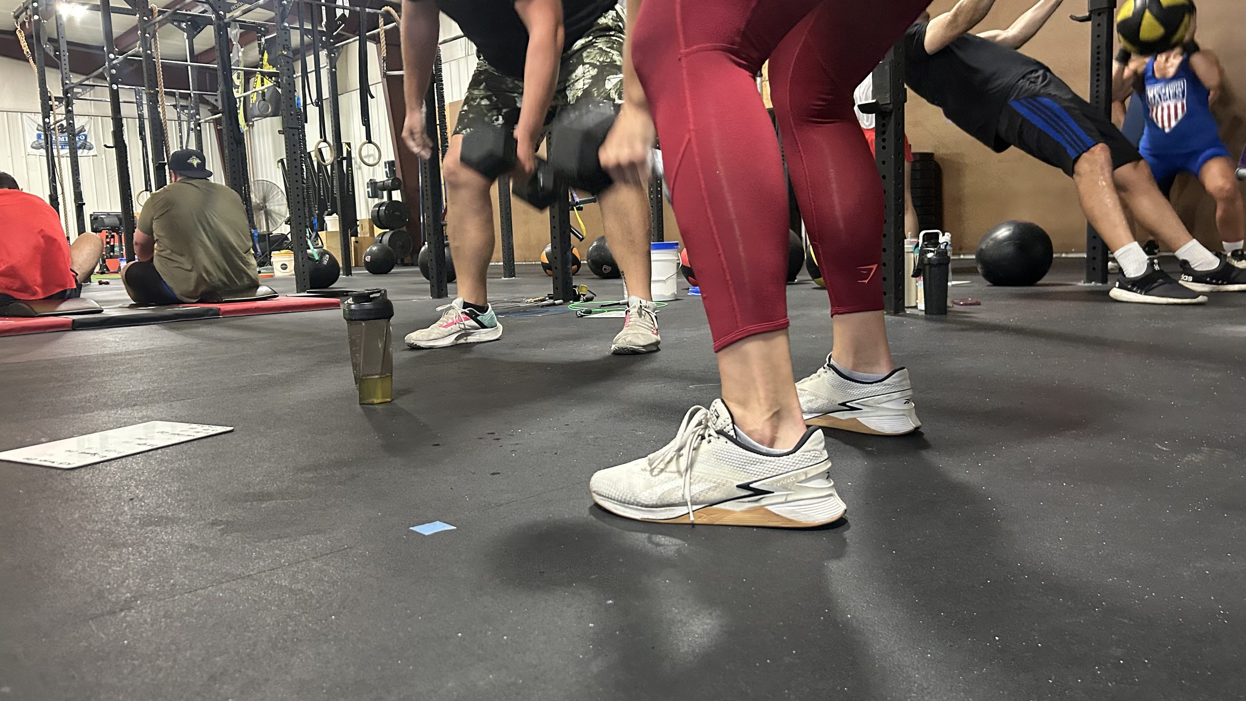 A floor-level view of one athlete's red leggings and white trainers mid-workout, with other athletes holding dumbbells and a medicine ball behind.