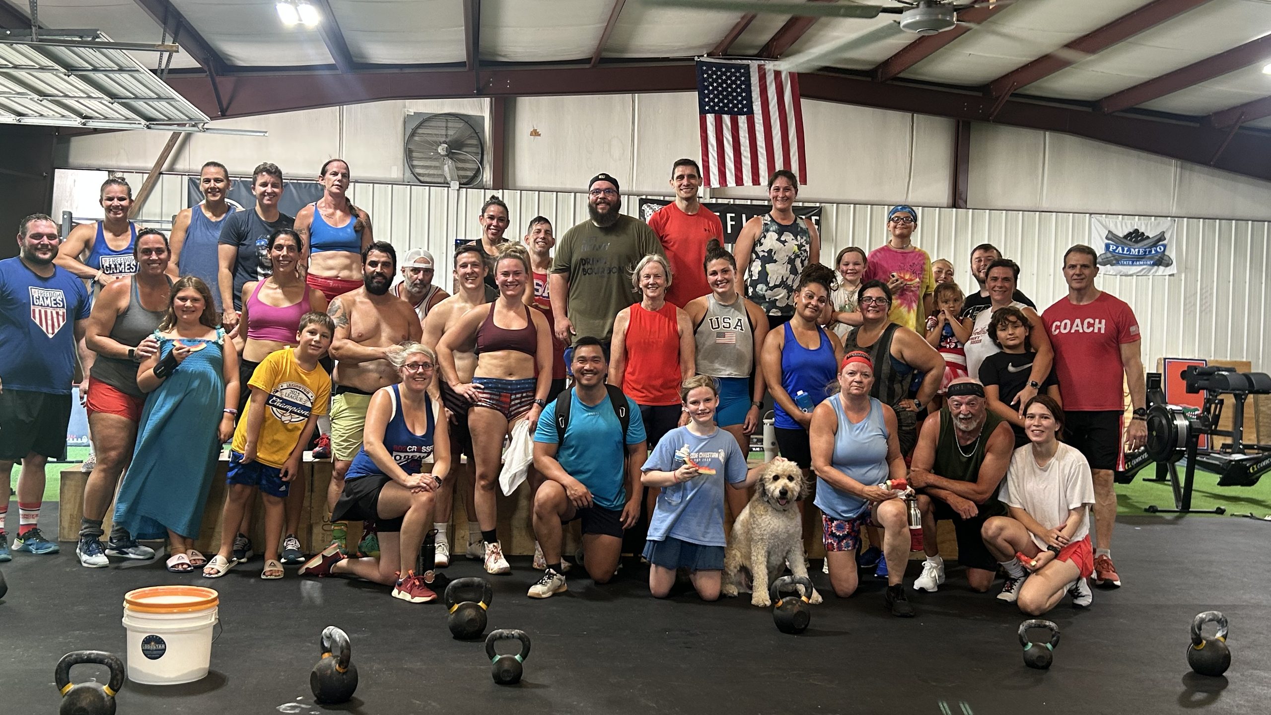 A large group of athletes, children and coaches gathers for a photo on the gym floor, with a cream-coloured doodle seated in the front row.