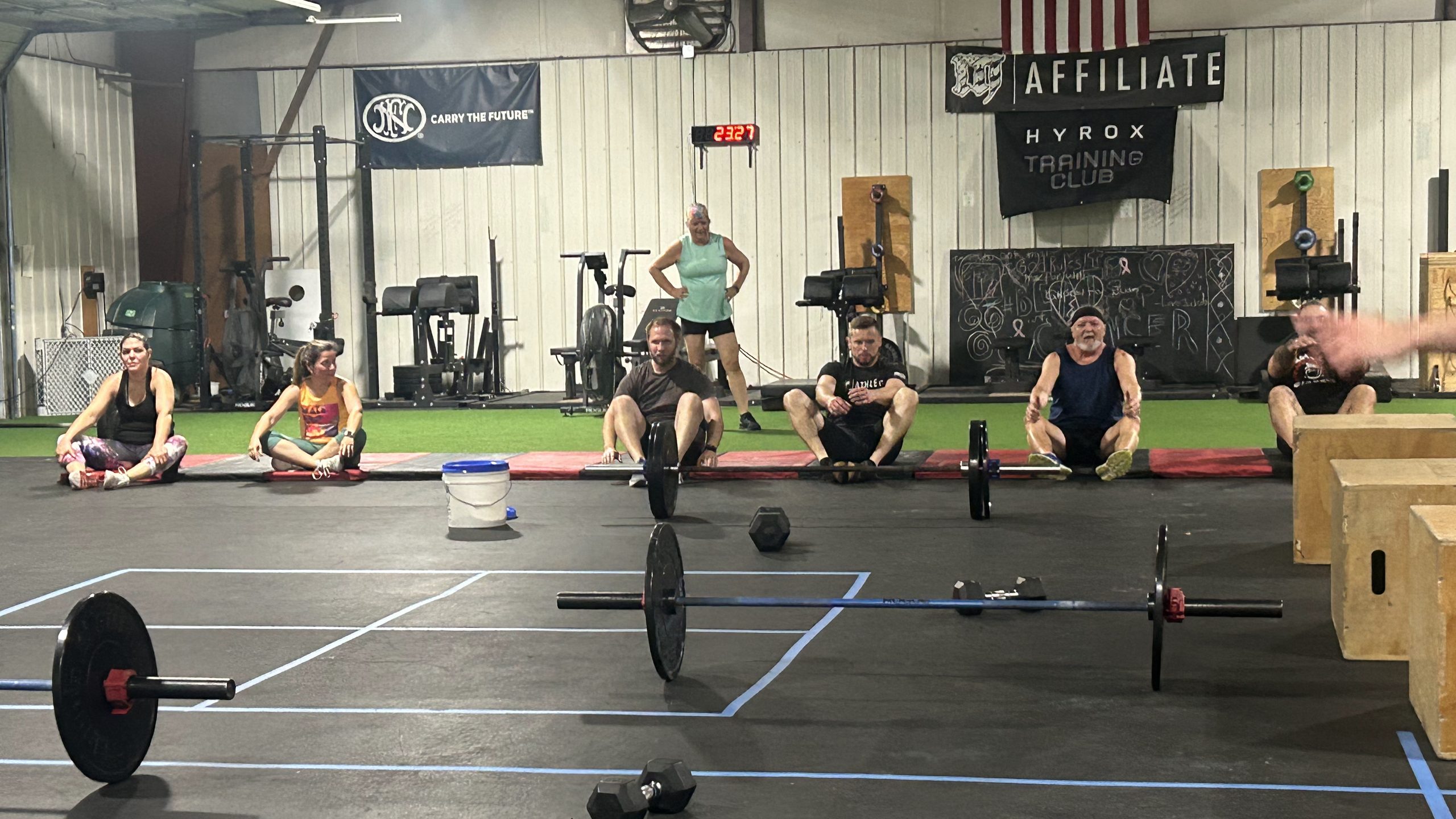 Athletes sit on the gym floor behind loaded barbells, with another athlete standing behind them.