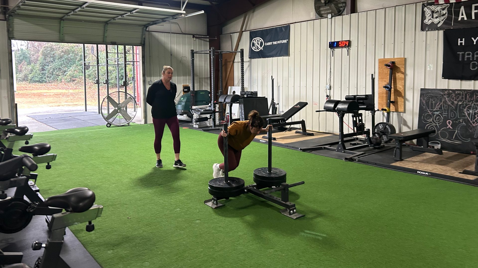 A coach watching a member push a loaded sled across the turf at Lodestar